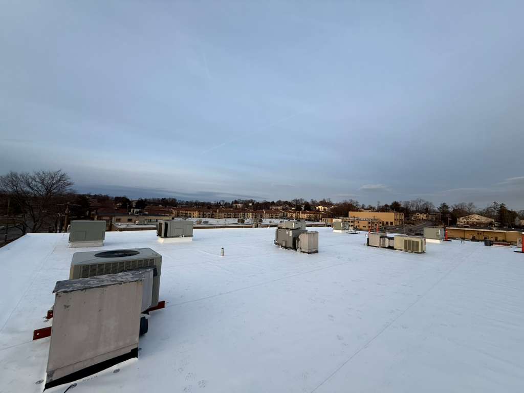 Snow-covered flat roof with multiple rooftop HVAC units and distant buildings.