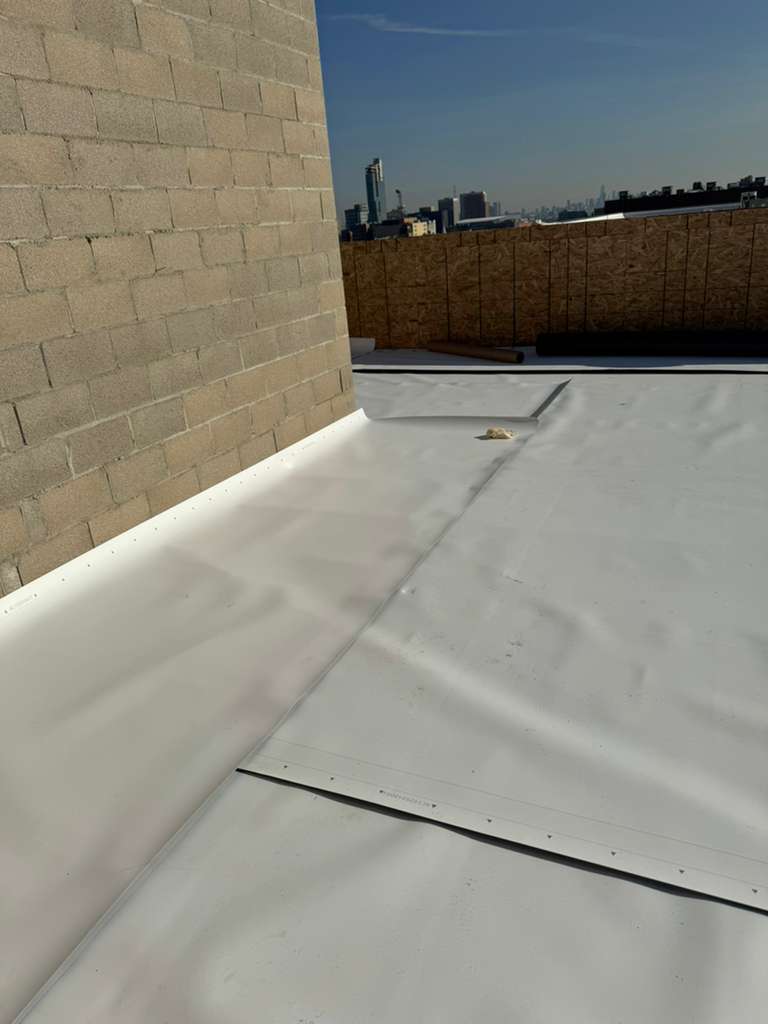 Flat white roof membrane during roof replacement on a rooftop with brick wall and distant skyline, New Jersey.
