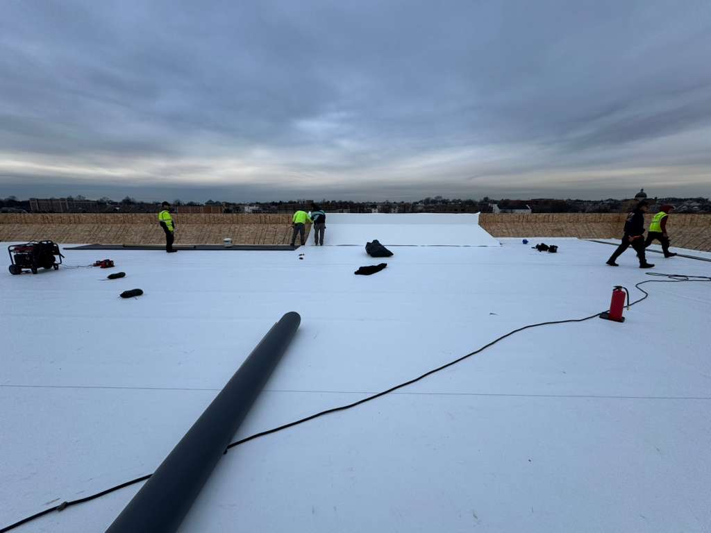 BlueSky roofing crew in high-visibility vests on a flat white roof performing roof replacement with rolled membrane and tools.