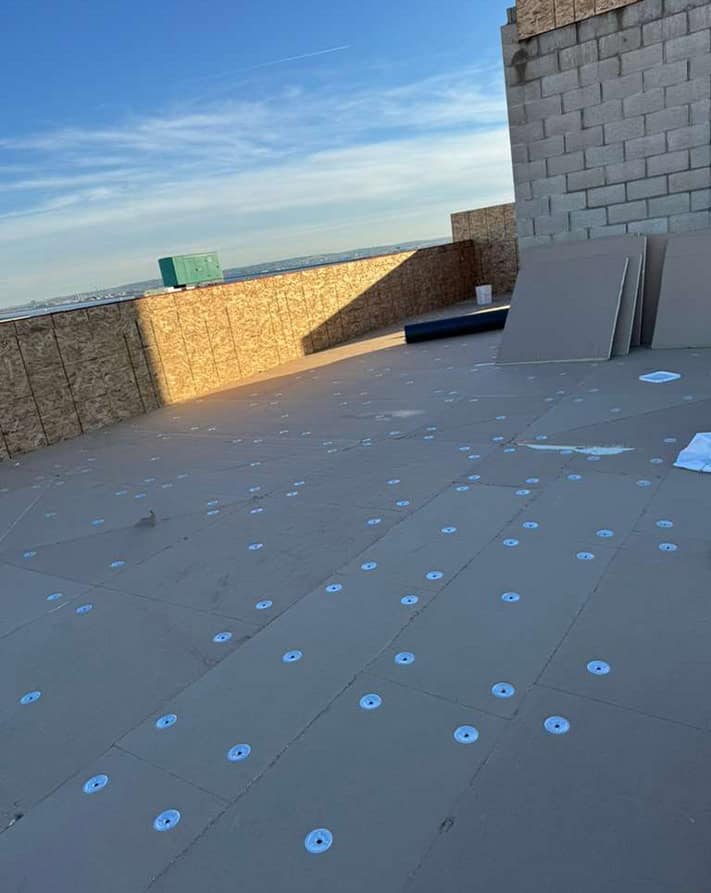 Flat roof under construction with gray membrane, circular fasteners, and nearby plywood panels.