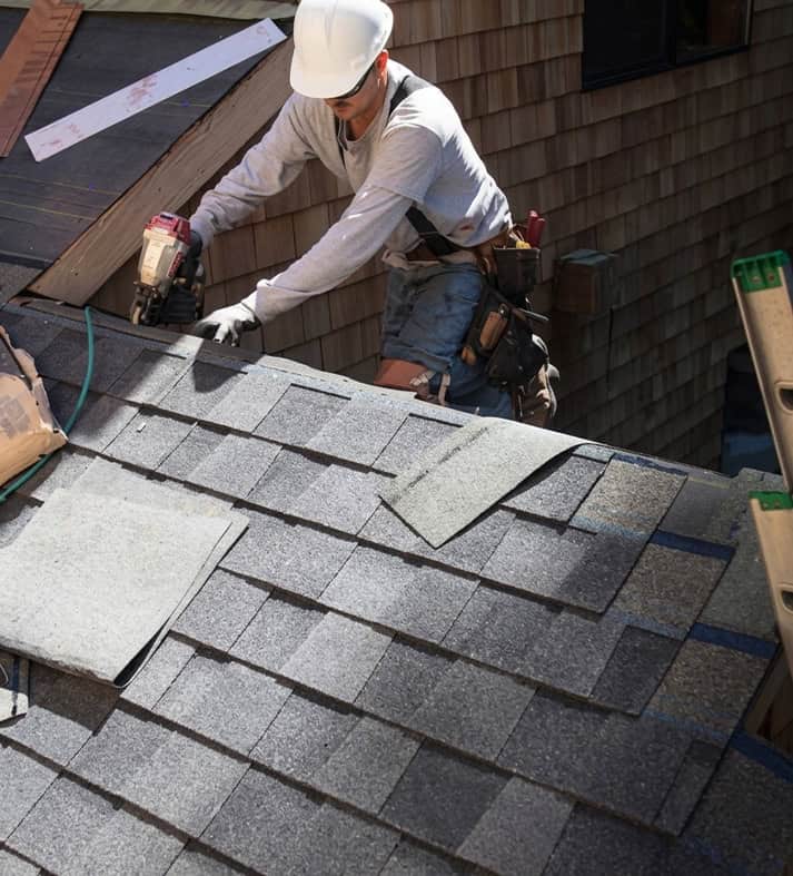 Worker wearing a white hard hat installs gray asphalt shingles on a sloped roof during a roof replacement.