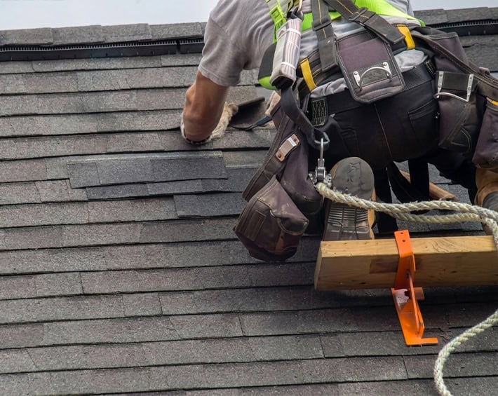 Roofer on dark asphalt shingle roof wearing safety harness during roof replacement.