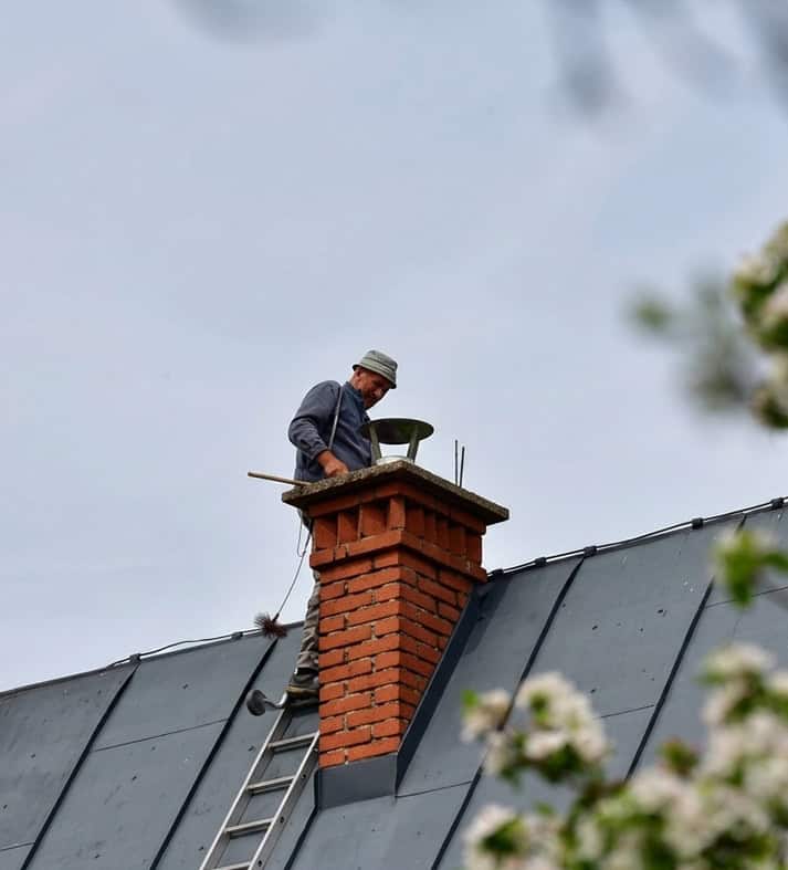 Roofer on ladder atop brick chimney performing chimney repair on a metal roof