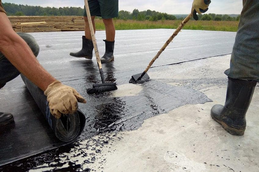 Workers lay black roofing underlayment on a flat roof with brushes and rollers.