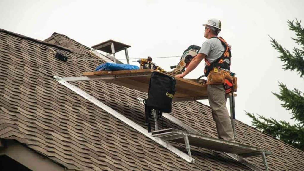 Roofer wearing helmet and safety harness atop brown shingle roof performing roof replacement in NJ