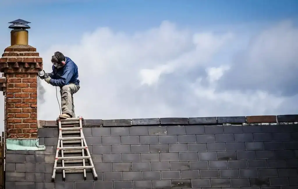 Worker on ladder performing chimney repair on brick chimney above slate roof.