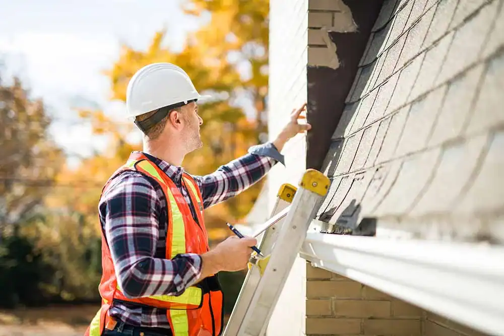 Worker in white hard hat and orange safety vest on ladder inspecting gutter installation along brick house in New Jersey.