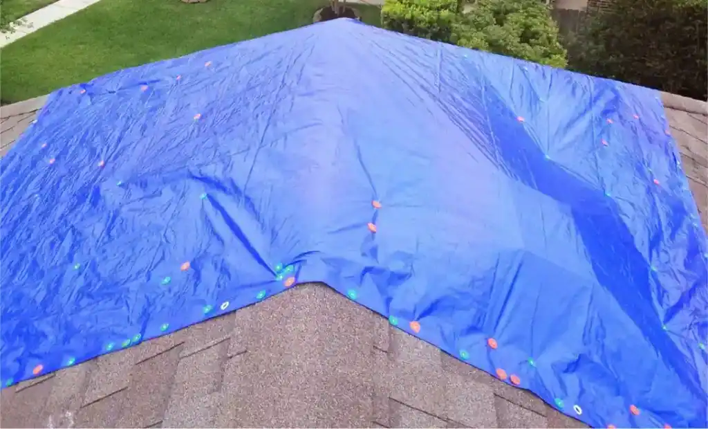Blue tarp covers a pitched asphalt shingle roof, orange and green fasteners securing it; emergency roofing in progress.