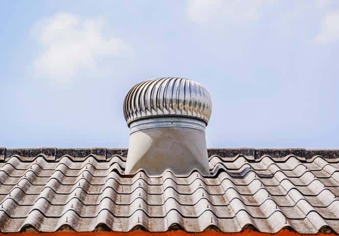 Ribbed metal vent cap atop a wavy tile roof under a blue sky.