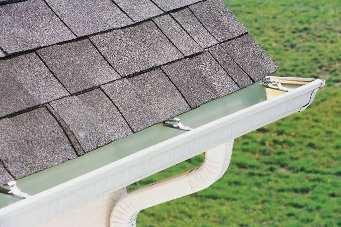 Close-up of asphalt shingle roof edge with white gutter installation and downspout brackets.