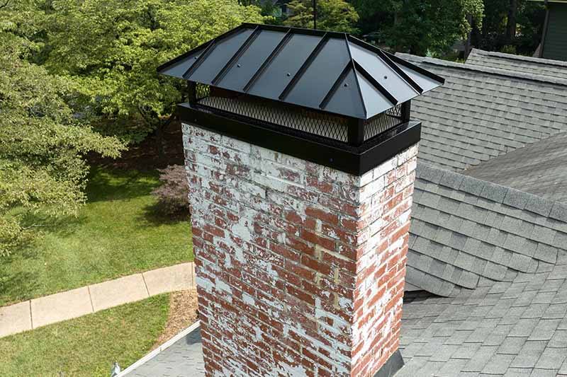 Weathered brick chimney masonry work with black metal cap on shingled roof in New Jersey