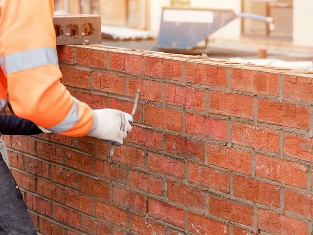 Worker in orange safety gear performing masonry work on a red brick wall.