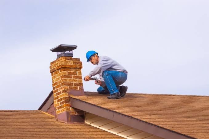 Blue-hatted worker on brown shingle roof performing chimney repair on brick chimney.